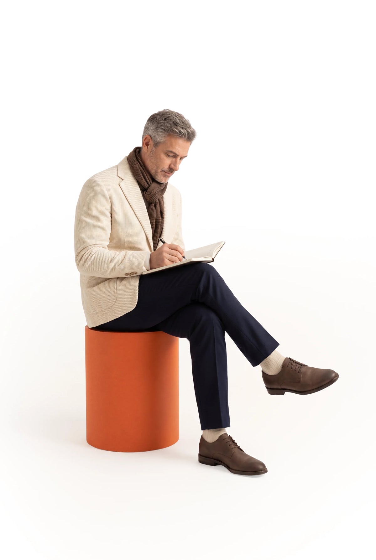 Man sitting on a small orange stool writing in a notebook on a white background