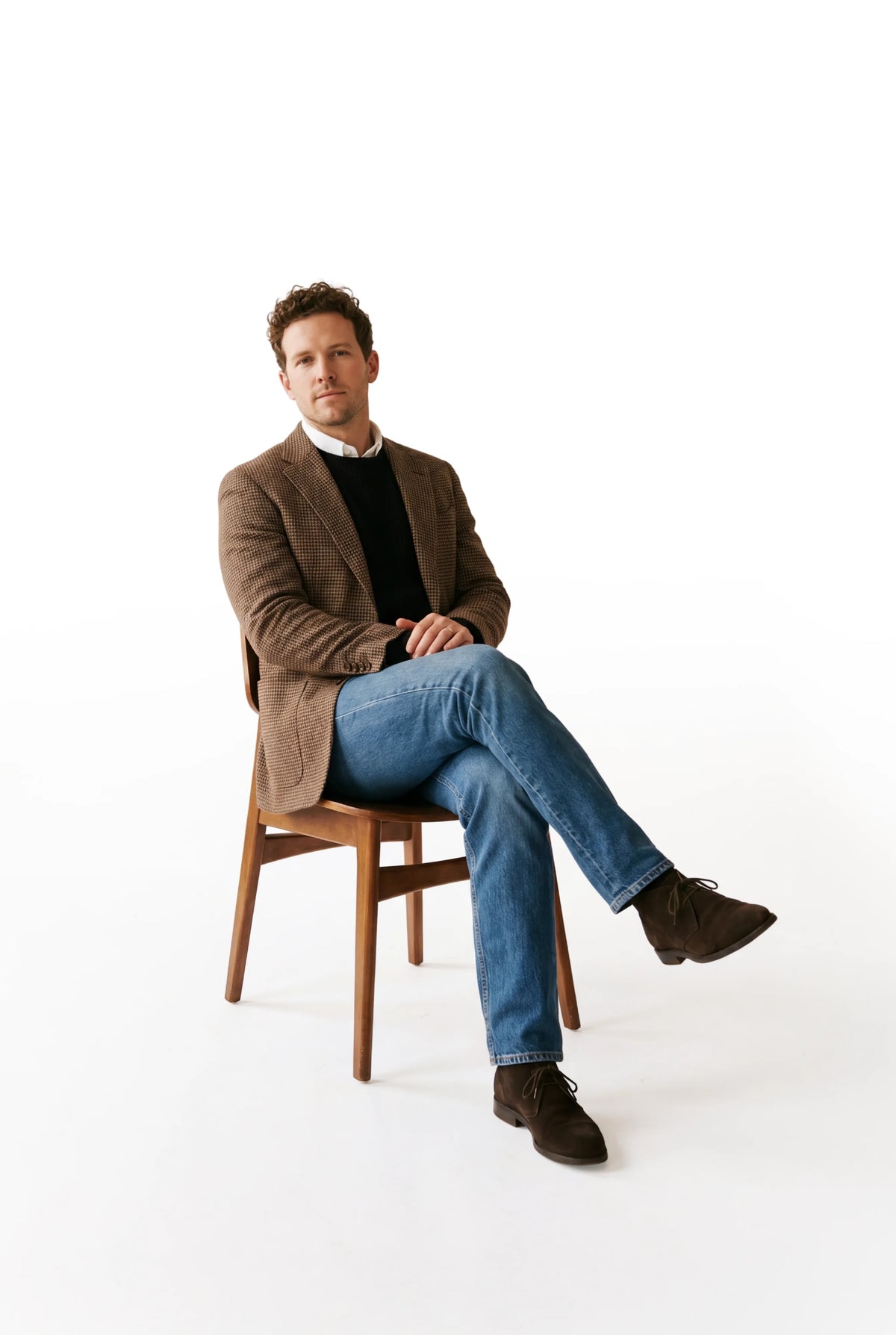 Man sitting on a wooden chair wearing a brown jacket, black shirt, blue jeans, and brown shoes on a white background.