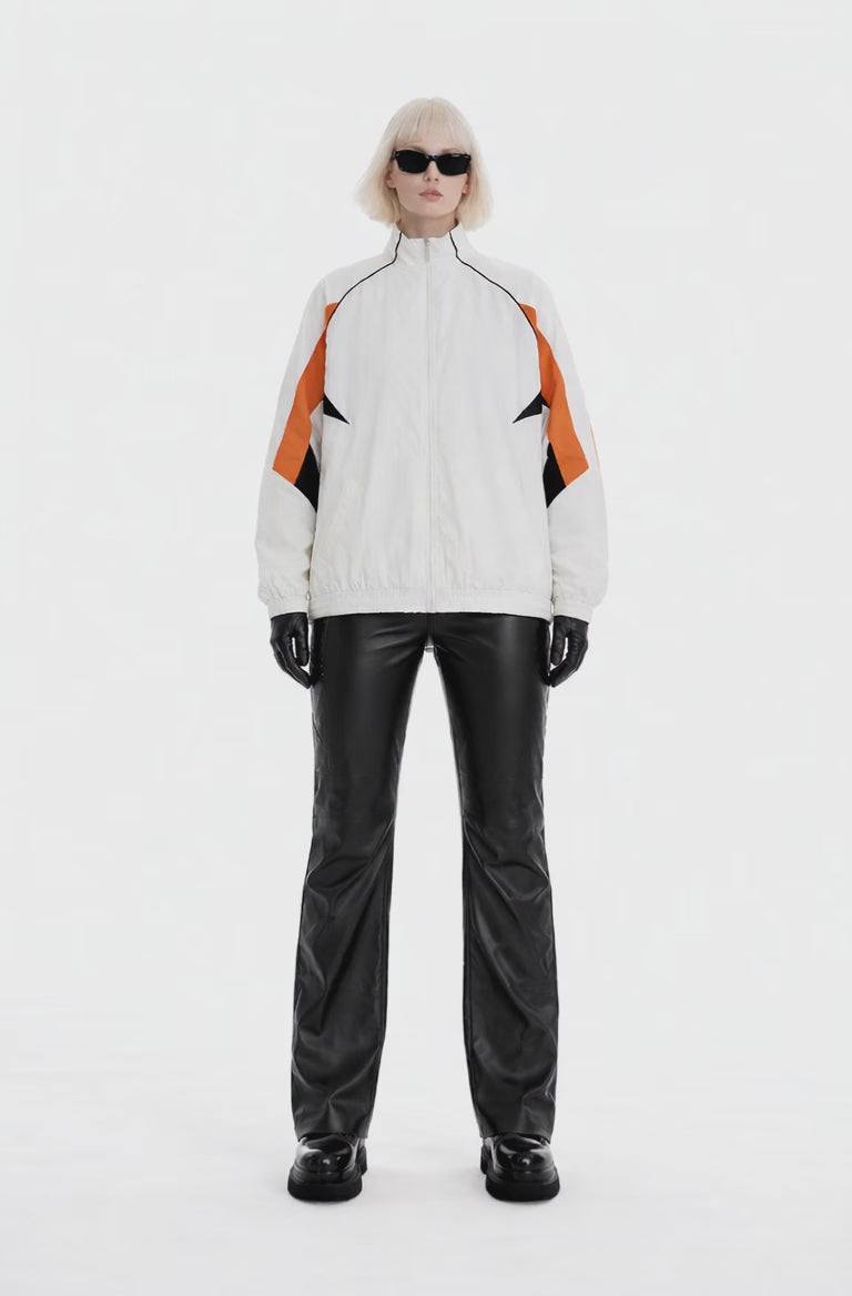 A full-body studio photograph of a woman wearing a white, athletic zip-up tracksuit jacket with orange and black geometric panels on the sleeves.