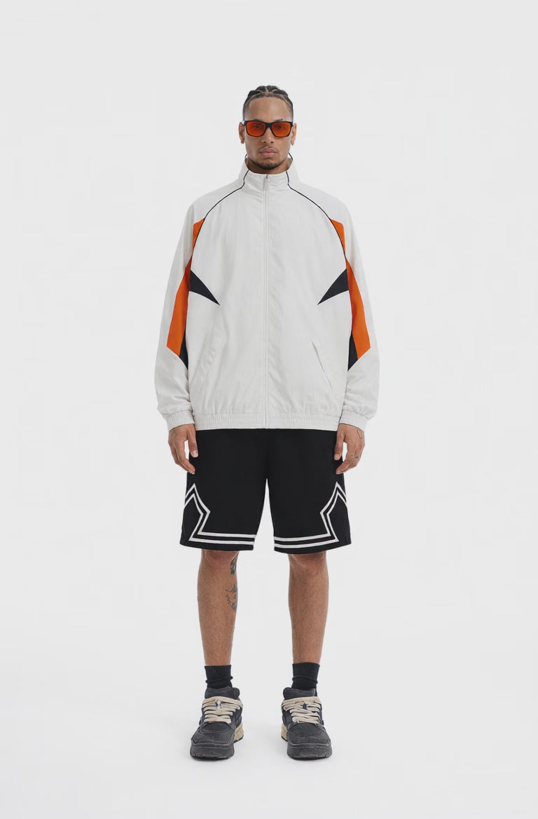 A full-body studio photograph of a man wearing a white, athletic zip-up tracksuit jacket with orange and black geometric panels on the sleeves.