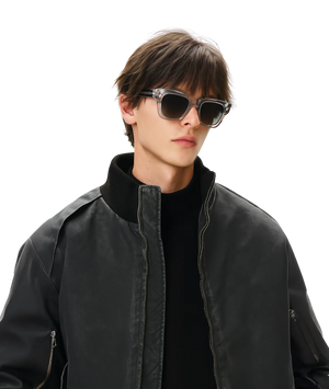 A man wearing stylish cat-eye sunglasses with a glossy black frame, a black turtleneck, and a black leather bomber jacket, set against a solid white background.