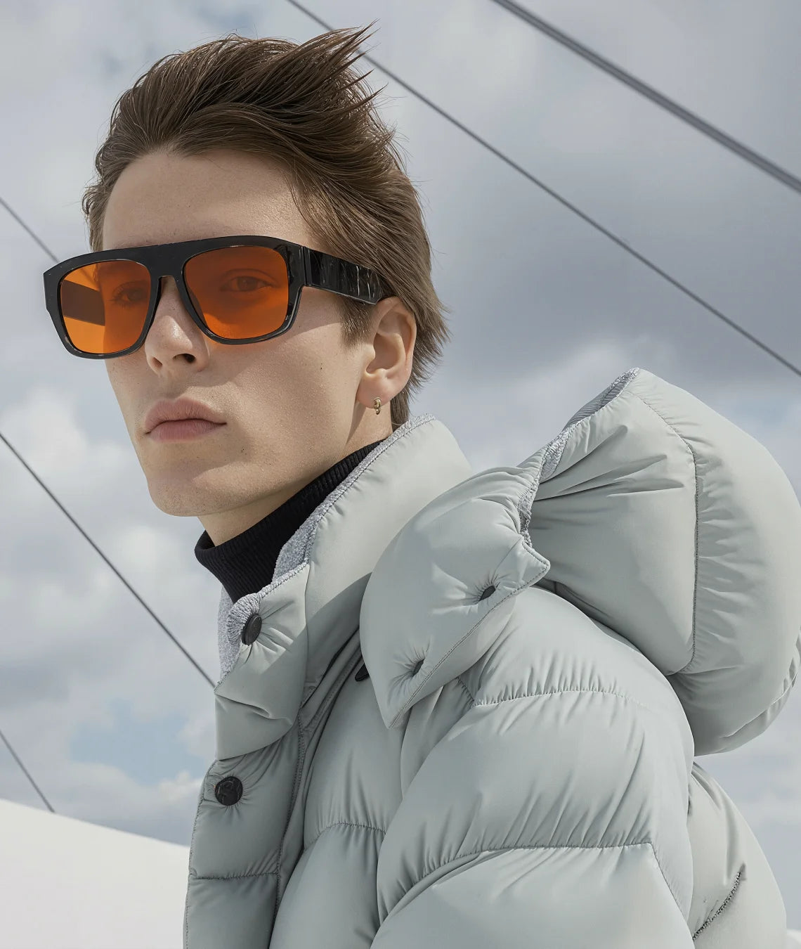 A close-up of a model wearing oversized square sunglasses with orange-tinted lenses and black frames, paired with a light gray puffer jacket and a black turtleneck, set against a cloudy sky with diagonal cable lines.